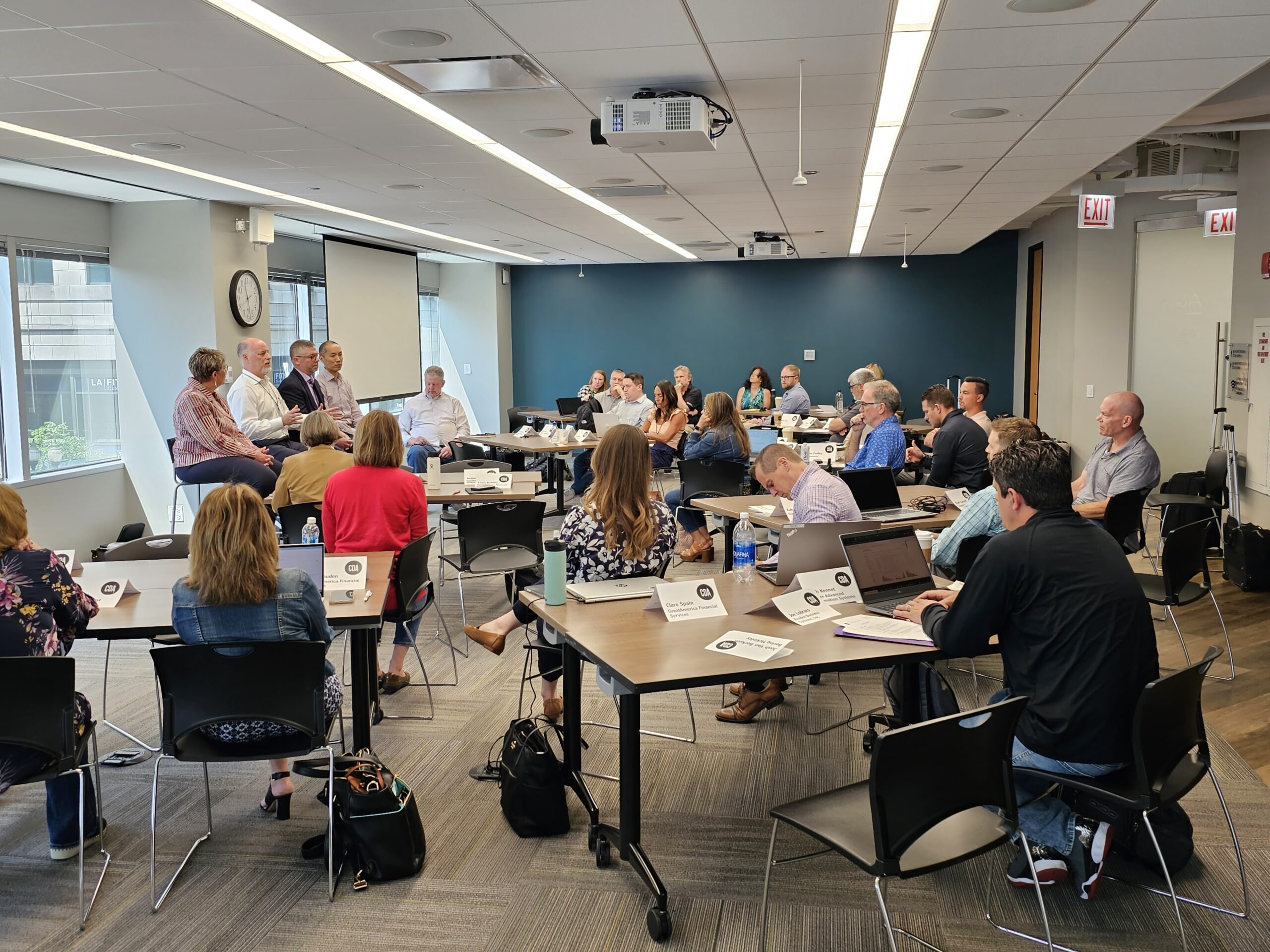 A roundtable discussion in a conference setting, with participants engaging in conversation and using laptops while seated at circular tables.