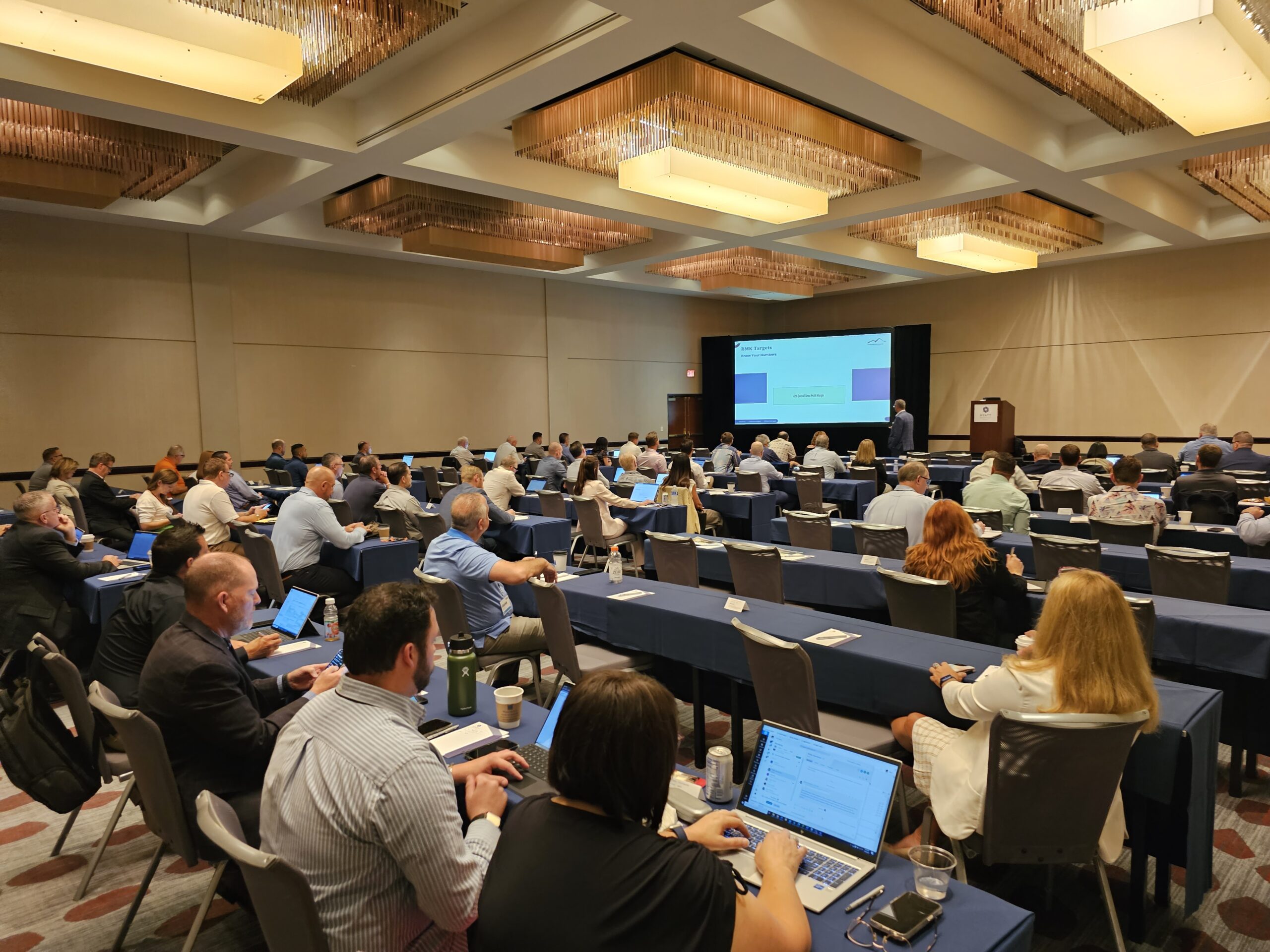 A large business conference with attendees seated at long tables, listening to a speaker at the front.