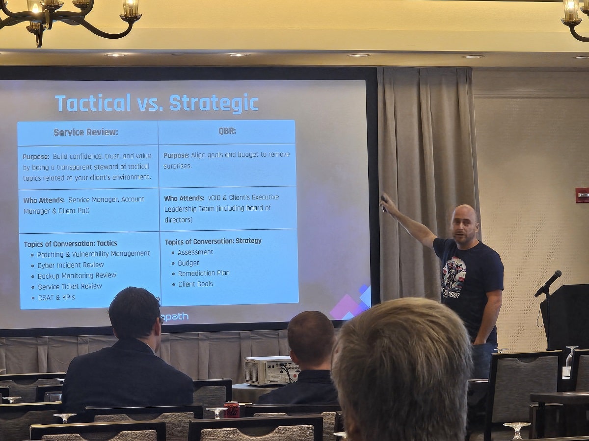 A man in a dark t-shirt presenting a slide titled "Tactical vs. Strategic" to an audience in a conference room.