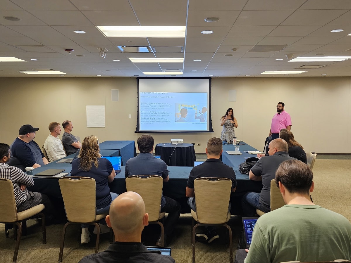 A conference room setting where a woman and a man are presenting to a group of attendees seated around a table with laptops.